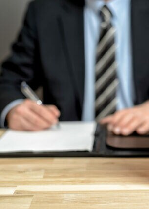 Eine Person im Anzug sitzt am Schreibtisch und unterzeichnet etwas, vor ihr stehen Gesetzesbücher.