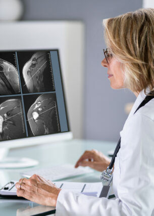 Eine Ärztin sitzt an ihrem Schreibtisch vor zwei Bildschirmen und kommuniziert mit einem Patienten via Videotelefonie.
