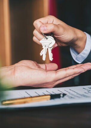 Eine Person hält einen Schlüsselbund über die aufgehaltene Hand einer anderen Person, darunter liegen ein Vertrag und ein Füller.