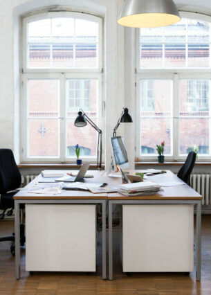 Zwei sich gegenüber stehende Schreibtische mit Unterlagen, PCs und Lampen sowie Regale vor zwei Fenstern.
