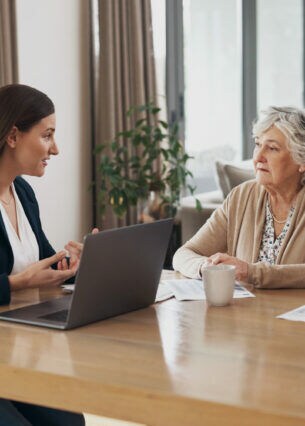 Ein älterer Mann und eine ältere Frau sitzen an einem Tisch. Vor ihnen liegen Dokumente. Gegenüber sitzt eine Frau vor einem Laptop.