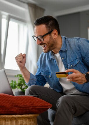 Ein Mann sitzt auf einem Sofa vor einem aufgeklappten Laptop, der auf einem Hocker steht. In einer Hand hält er eine Geldkarte, die andere ballt er zu einer Faust.