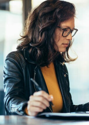 Eine Frau vor einem Laptop hält den Zeigefinger einer Hand auf das Laptop-Touchpad, in der anderen Hand hält sie einen Stift.
