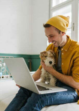 Eine Person sitzt auf einem Sofa, krault einen Hund auf dem Schoß und blickt in ein geöffnetes Notebook.