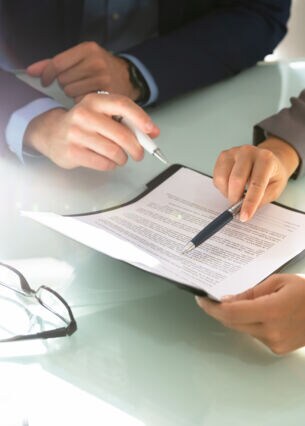 Zwei Personen an einem Tisch zeigen mit Finger und Stift auf ein Dokument in einem Klemmbrett.