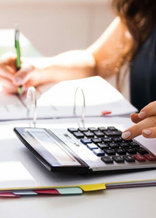 Eine Person tippt mit einer Hand auf einem Taschenrechner, der auf einem aufgeschlagenen Aktenordner liegt, in der anderen Hand hält sie einen Stift.