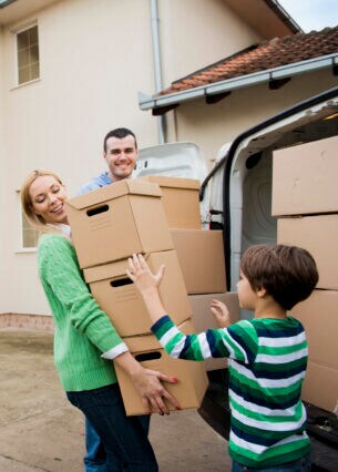 Ein Mann, eine Frau und ein Kind stehen Kisten tragend an einem offenen Transporter vor einem Haus.