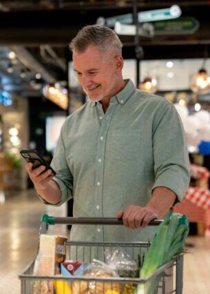 Eine Person steht mit einem Einkaufswagen im Supermarkt und schaut auf ihr Handy in der Hand.