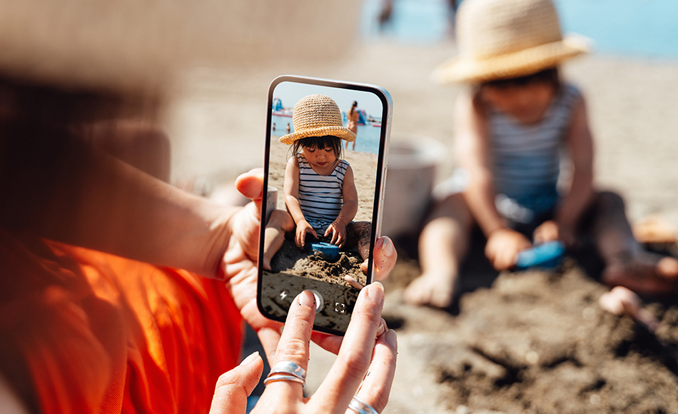 Ein Kind spielt am Strand und ein Elternteil nimmt ein Bild mit einem Smartphone auf.